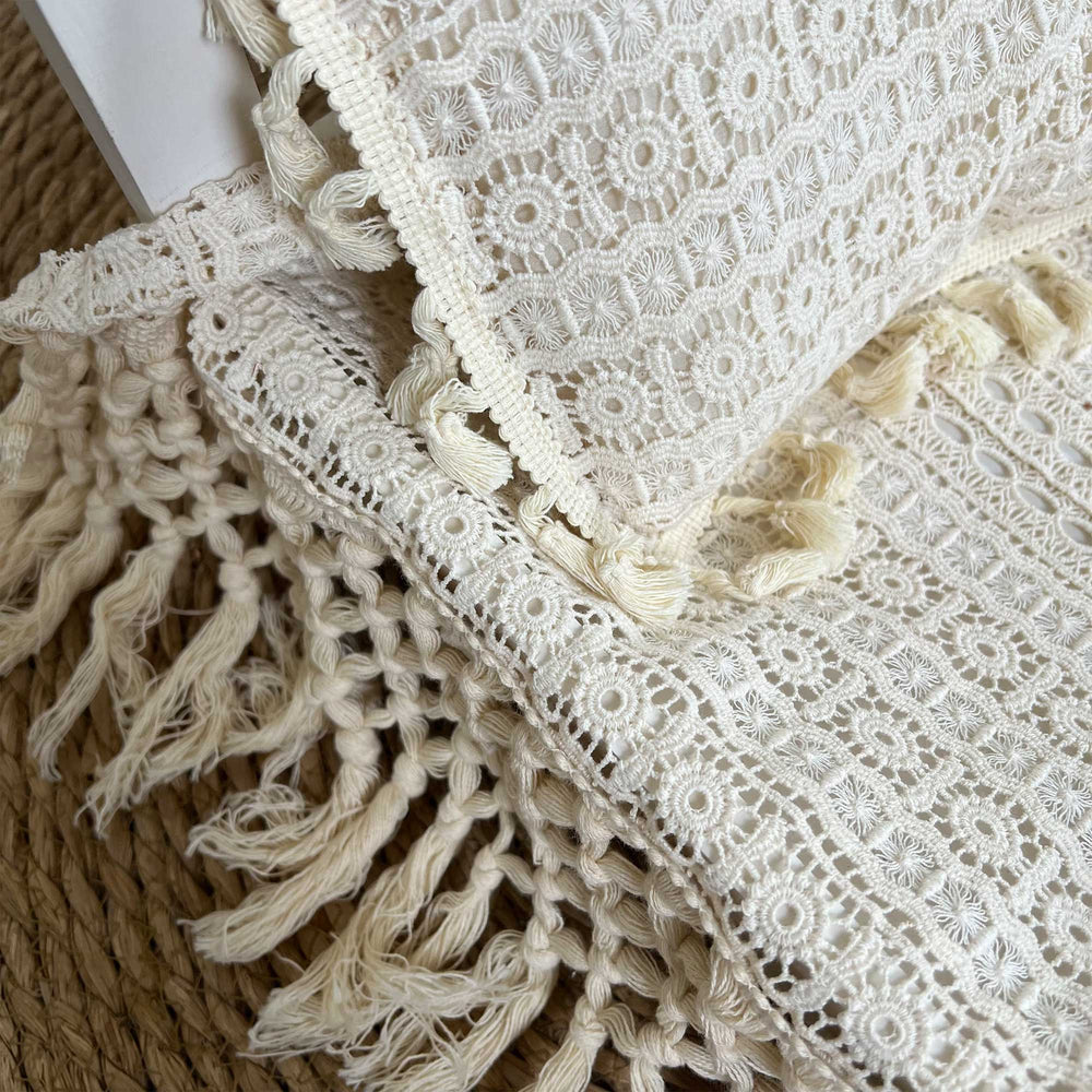 A close-up view of a beige cotton crochet textile set featuring a blanket with a geometric openwork pattern and long knotted fringe.