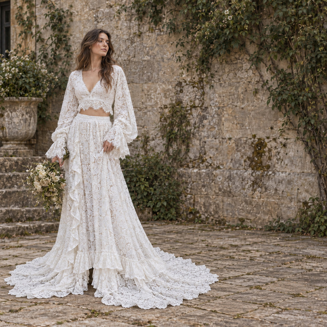 A woman stands outdoors in front of a stone wall and steps, wearing a ivory lace two-piece outfit with a long-sleeve cropped top and a full ruffled skirt with a train. 