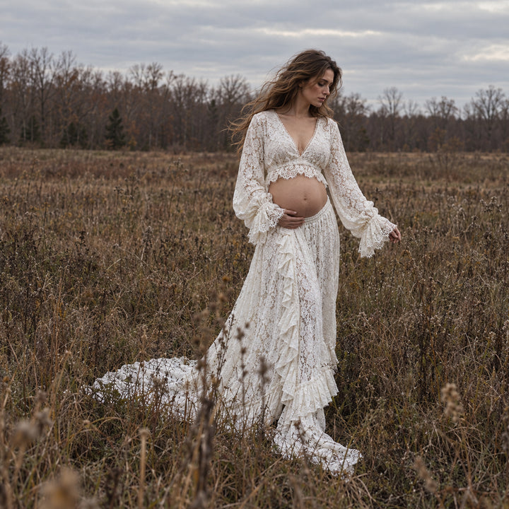 A pregnant woman stands in a dry grass field wearing a ivory lace bohemian-style maternity dress with a matching cropped top. The dress features long bell sleeves, a deep V-neckline, and a tiered ruffled skirt with a long train.