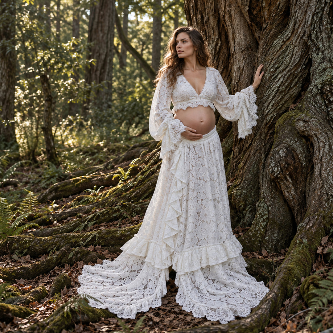 A pregnant woman wearing a ivory lace maternity gown stands in a forest next to a large tree trunk, holding her baby bump. The dress features a lace crop top with long ruffled sleeves and a matching long ruffled skirt with a train.