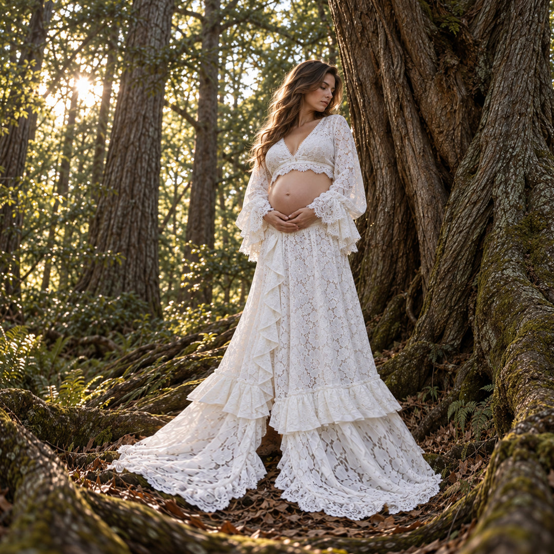 A woman wearing a ivory lace maternity dress stands outdoors in a forest beside a large tree trunk, holding her baby bump. The dress has a deep V-neck, long ruffled sleeves, and a tiered skirt with a flared hem.