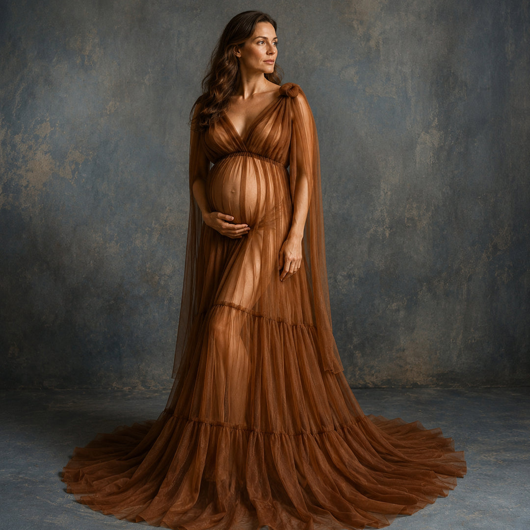 A pregnant woman stands in a studio wearing a sheer, floor-length brown gown with a draped cape-like overlay and a full skirt that spreads across the floor. 
