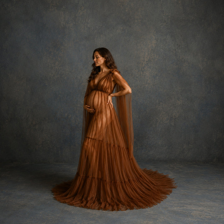 A pregnant woman stands in profile wearing a flowing brown gown with a sheer draped cape and a full layered skirt.