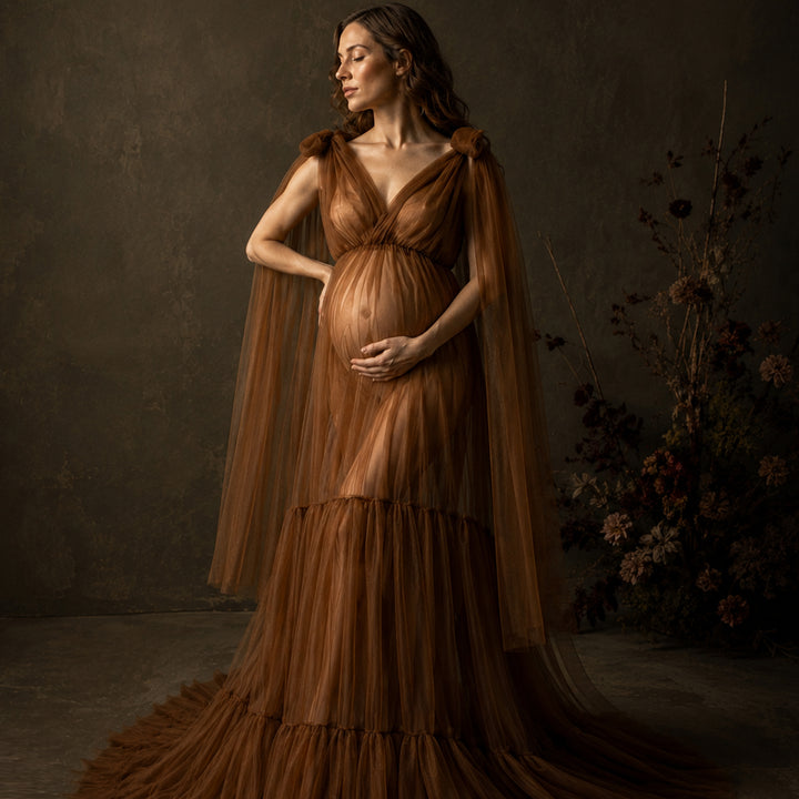 A pregnant woman wearing a luxurious brown tulle gown with a draped cape-style overlay, standing in a dark, moody studio setting with dried floral arrangements in the background.