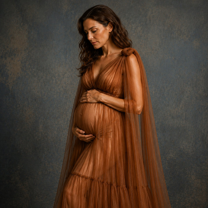 A pregnant woman wearing a flowing brown maternity gown with a sheer draped cape, standing against a textured dark gray studio backdrop.