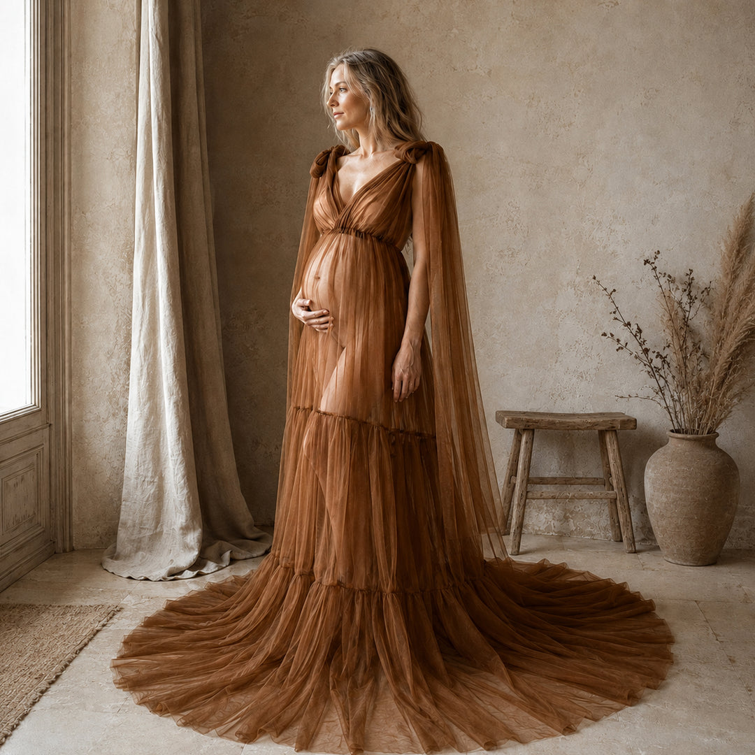 A woman stands in a rustic room wearing a floor-length brown tulle gown with a draped cape-style overlay and a full skirt that spreads across the floor. 
