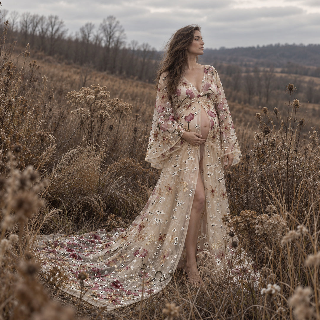 A pregnant woman stands in a dry grassy field wearing a long, sheer tulle gown with floral embroidery and sequined detailing.