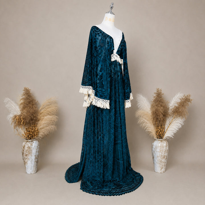 A long green velvet dress displayed on a mannequin against a warm beige studio backdrop, flanked by two vases of dried pampas grass. The dress has a deep V-neck, long sleeves with white lace trim at the cuffs, and a matching headpiece.