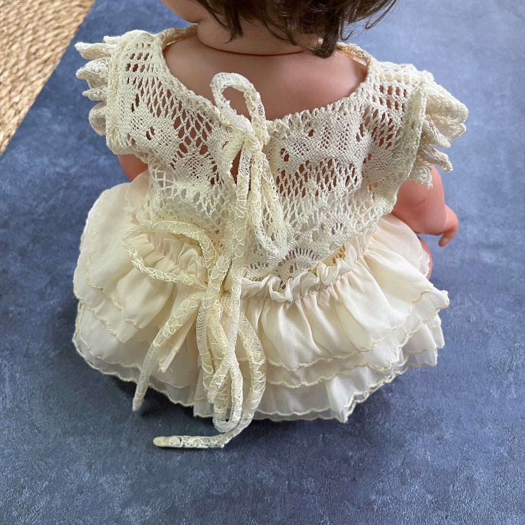 The doll wears a beige crocheted lace romper with a back tie closure and a double layer ruffled skirt hem.
