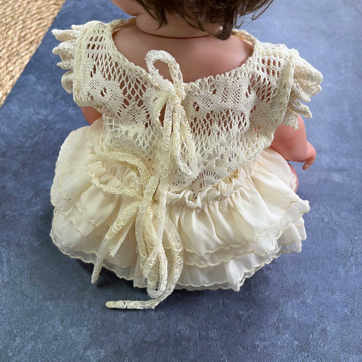 The doll wears a beige crocheted lace romper with a back tie closure and a double layer ruffled skirt hem.