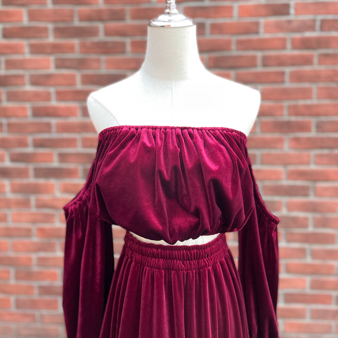 A deep burgundy velvet off-the-shoulder crop top and matching skirt set displayed on a white dress form mannequin, set against a red brick wall background.