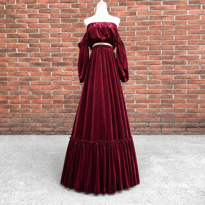 A floor-length off-the-shoulder velvet gown in deep burgundy red, displayed on a white mannequin torso against a red brick wall and concrete floor.