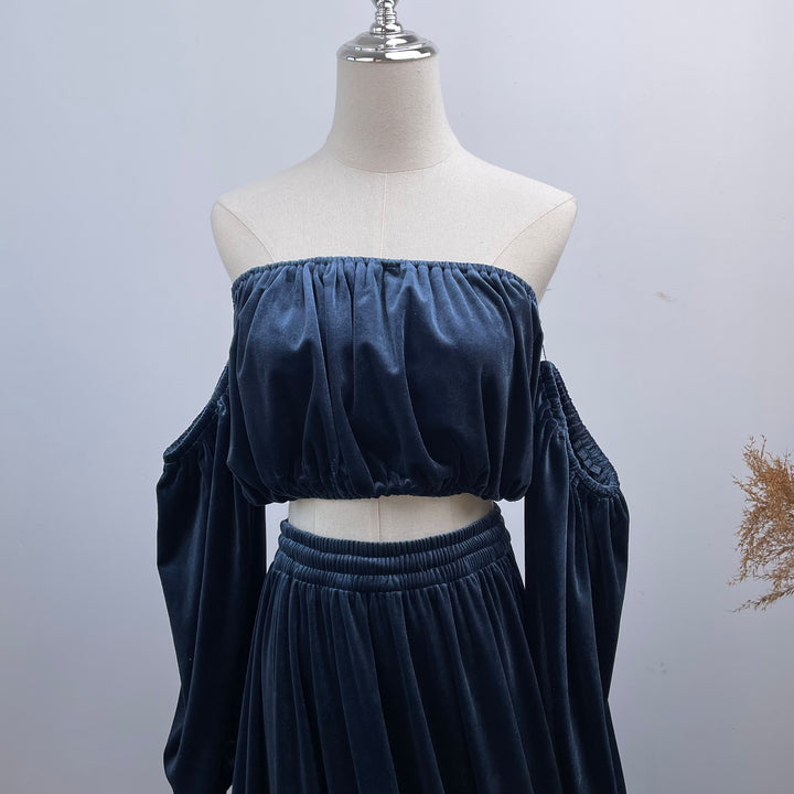 A dark navy blue velvet two-piece set displayed on a cream mannequin torso. The set includes an off-the-shoulder crop top with long draped sleeves and a matching high-waisted maxi skirt with an elastic waistband.