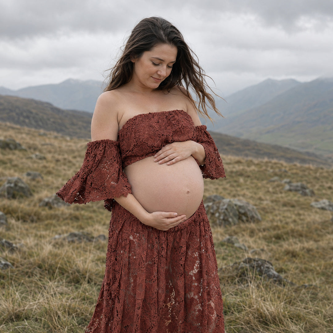 A pregnant woman stands in profile on a misty, grassy hillside wearing a terracotta two-piece lace maternity set with off-the-shoulder ruffled sleeves and a high-waisted skirt that reveals her bare baby bump against a mountain backdrop.