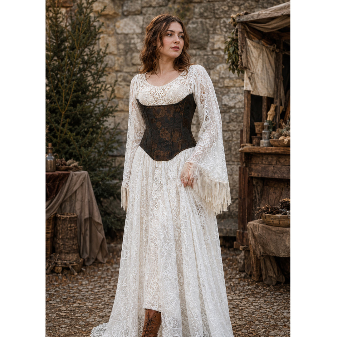 A woman stands outdoors in a rustic stone setting wearing a long white lace dress with a fitted dark brown corset-style bodice, sheer long sleeves with fringe, and a full skirt.