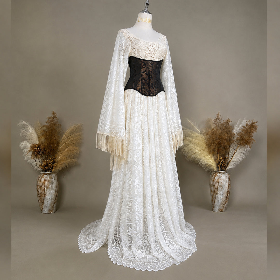 A long beige lace dress displayed on a mannequin against a warm taupe studio background, with a black lace corset bodice and two long sheer lace sleeves with fringe. Two vases with dried pampas grass are placed on either side.