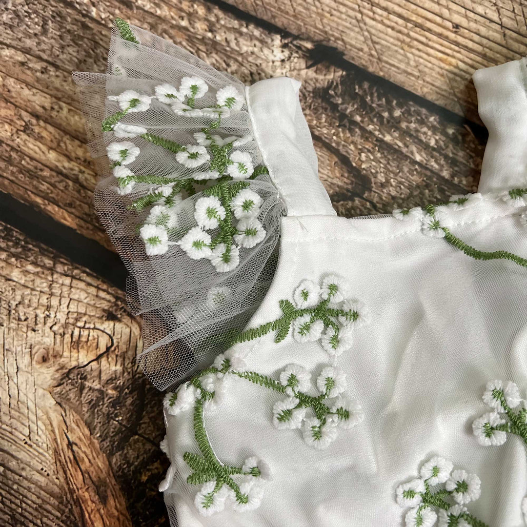 A close-up of the strap and bodice of the same romper, showing the tulle ruffle, white 3D flowers and green embroidered stems, on a wood-grain surface.