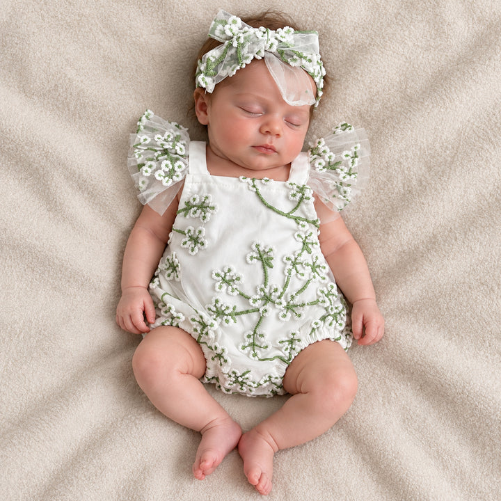 A baby lying on her back on a beige blanket with eyes closed, showing the embroidered front of the romper and the tulle flower straps on both sides.