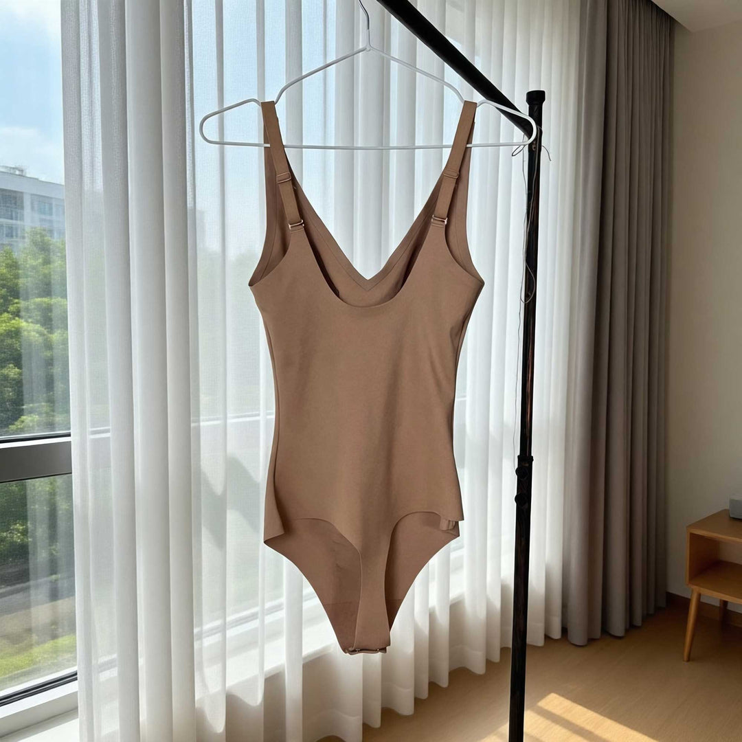 A khaki Callirrhoe bodysuit is hanging on a hanger in front of a curtain next to a window.
