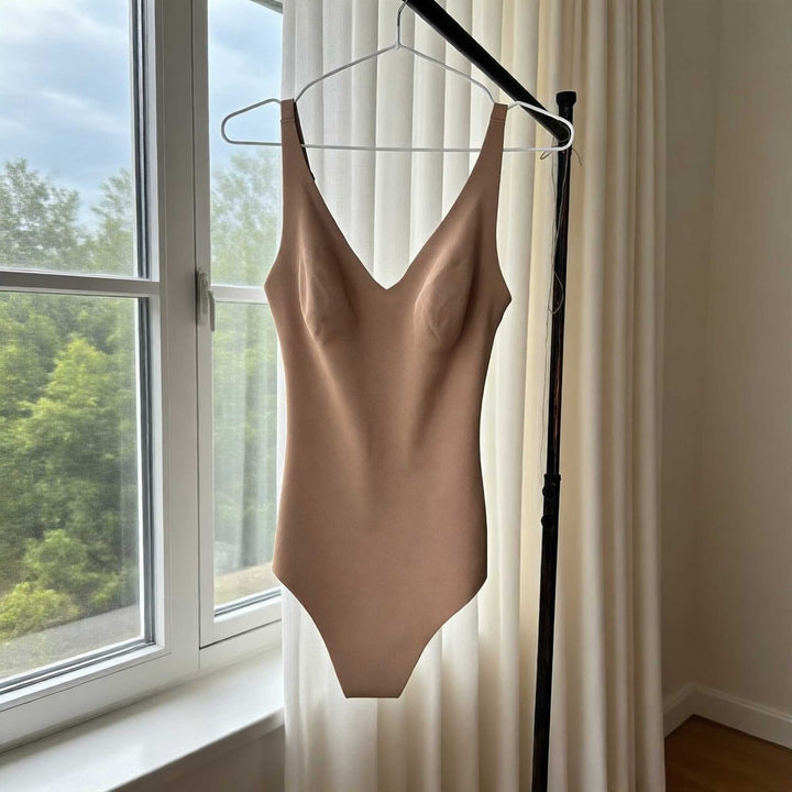 A khaki Callirrhoe bodysuit is hanging on a hanger in front of a curtain next to a window.