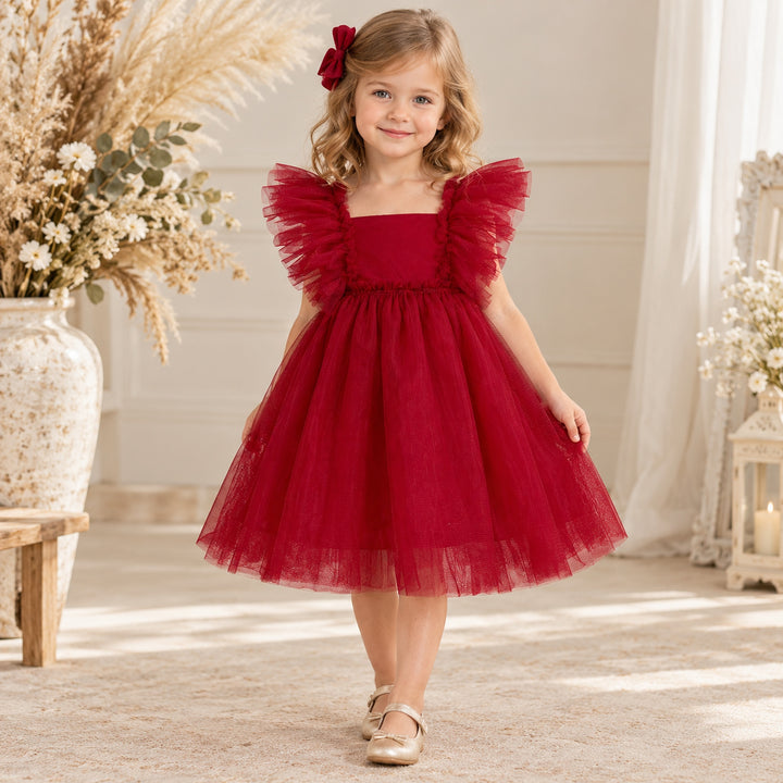 A girl in a Callisto red tulle dress with a red bow headband and light shoes stands indoors beside flower arrangements.