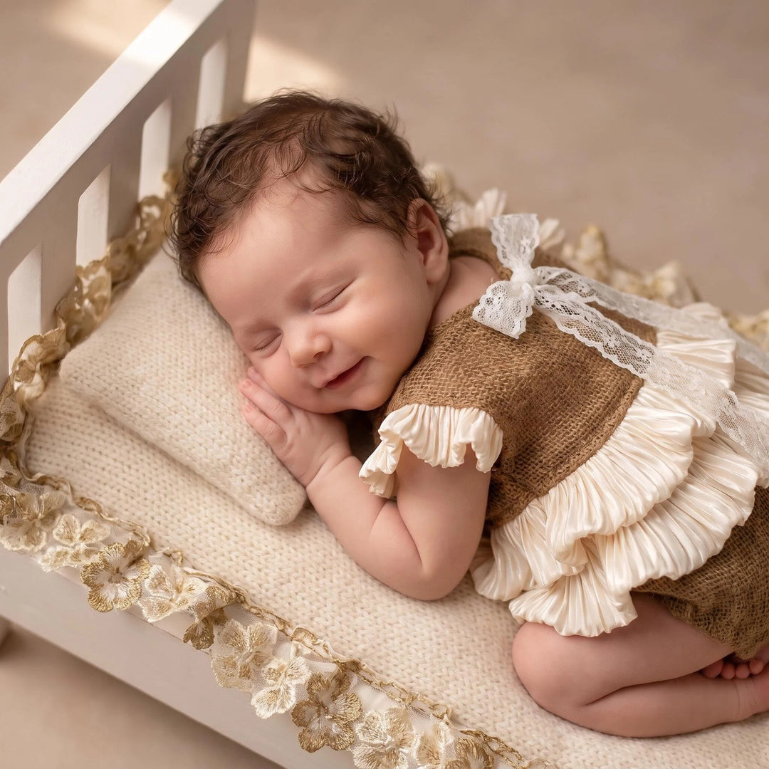 A newborn lies prone in a white wooden crib, wearing a camel mohair knit romper with double-layer beige crystal satin ruffles and white lace shoulder straps.