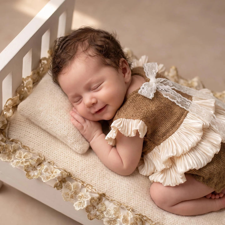 A newborn lies prone in a white wooden crib, wearing a camel mohair knit romper with double-layer beige crystal satin ruffles and white lace shoulder straps.