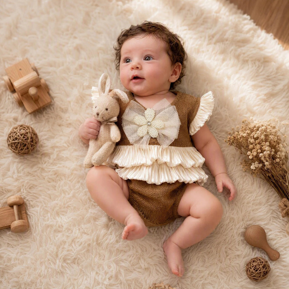 A newborn lies supine on a beige plush rug, wearing a camel knit romper with a mesh bow and double-layer ruffles, holding a beige rabbit toy.