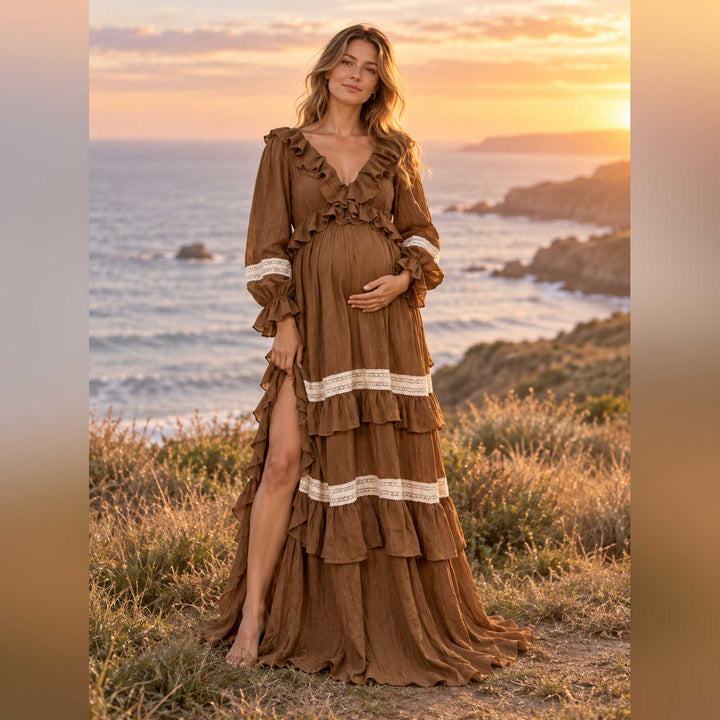A pregnant woman in the Cassandra coffee tiered dress stands barefoot on coastal grass, one hand on her belly, with sea and sunset behind.