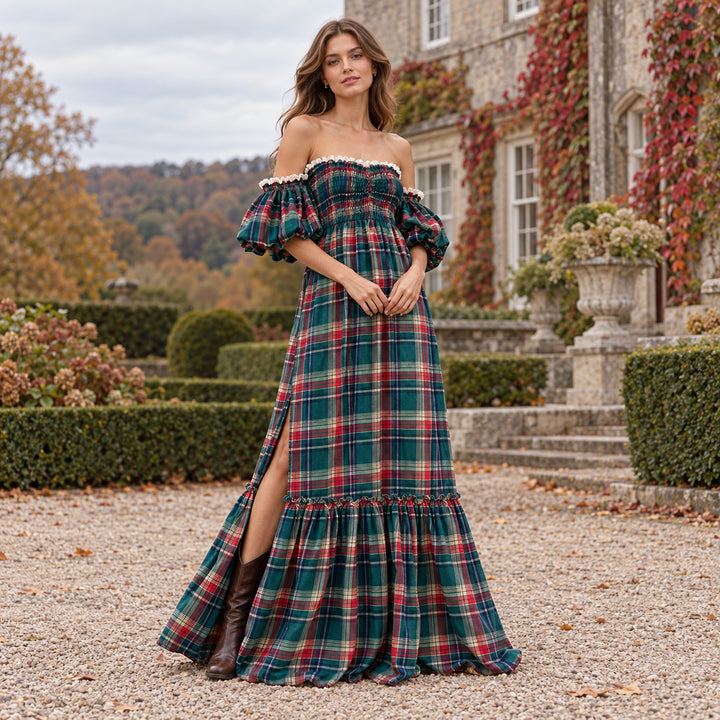 A woman in Catubri plaid off-shoulder maxi dress with brown knee-high boots stands on a gravel path, with a stone house, vines and shrubs behind.