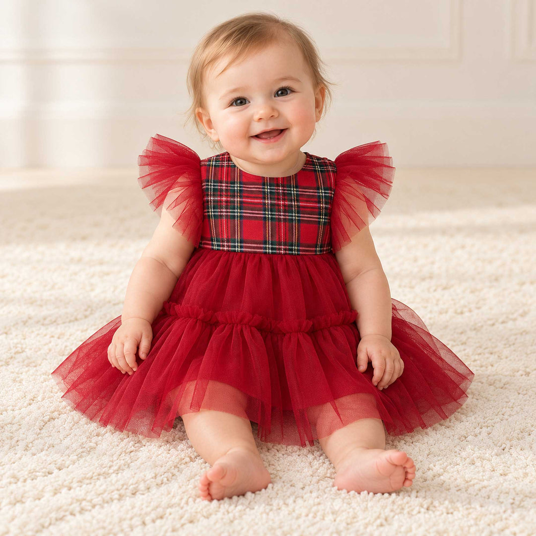 A baby in the Cerridwen dress sits on a light carpet facing the camera, with a white interior wall behind.