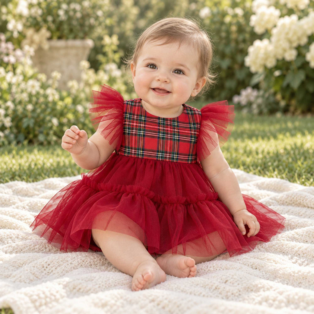 A baby in a Cerridwen plaid bodice dress with a red tulle skirt sits on an outdoor blanket, grass and flowers behind.