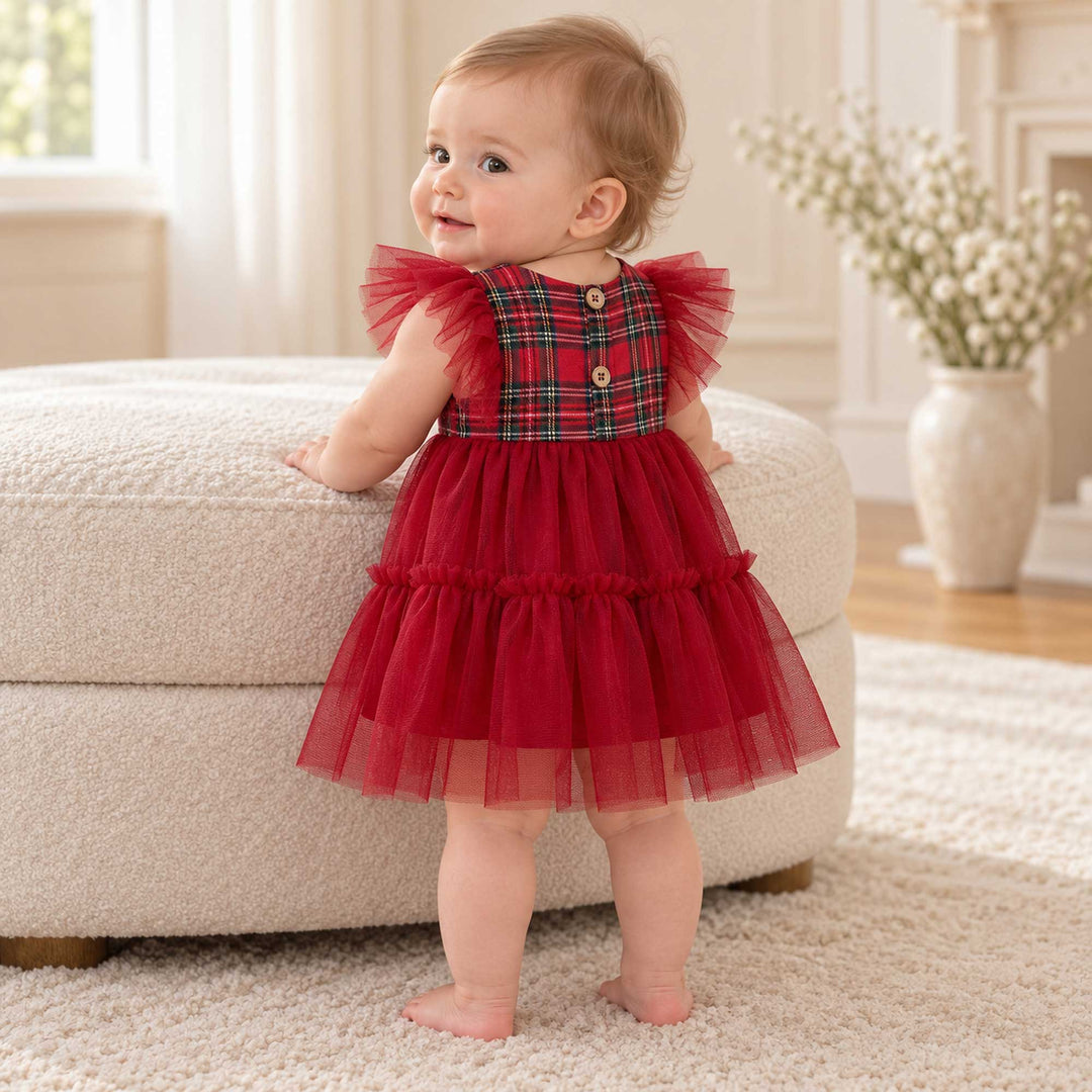 A baby in the Cerridwen dress stands with her back turned and head turned, one hand on an ottoman, buttons on the back, a vase on the right.