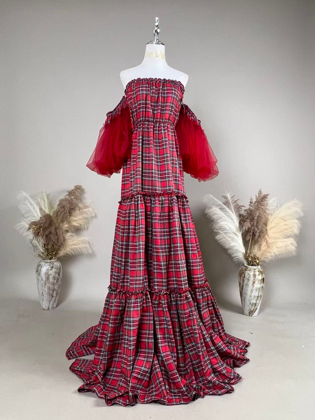 A Charybdis plaid off-shoulder dress with red tulle sleeves on a mannequin facing forward, tiered skirt, train on the floor, a dried-flower vase on each side.
