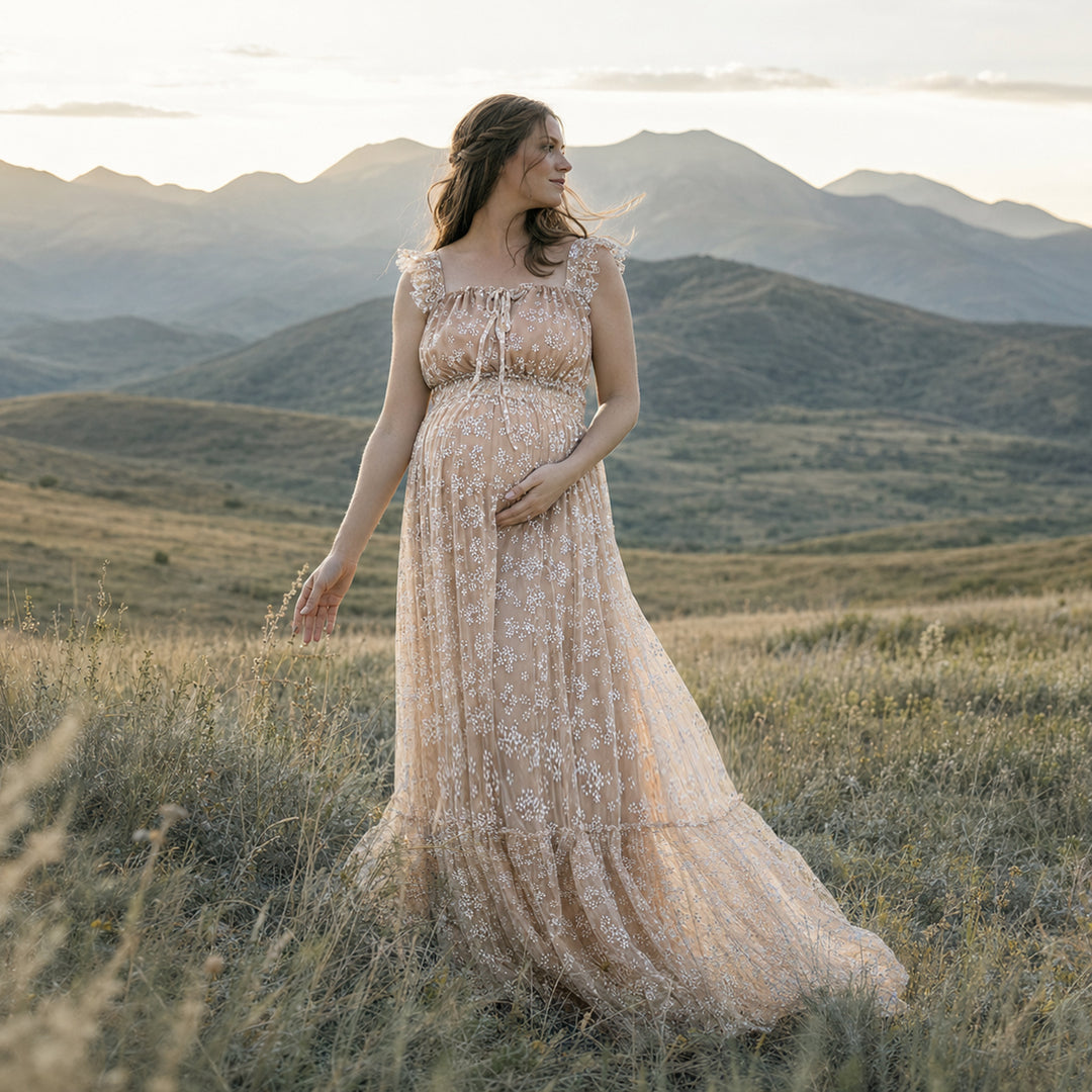 A pregnant woman stands in a grassy mountain landscape wearing a khaki floral embroidered tulle maternity gown with ruffled cap sleeves and a flowing skirt.