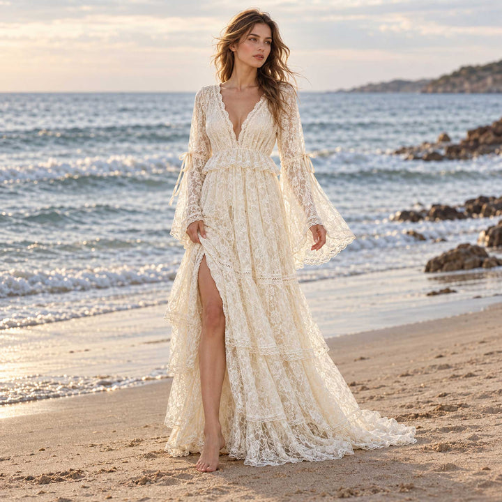 A woman in a Clito beige lace long-sleeve dress with a V neckline, side slit and tiered skirt stands barefoot on a beach, sea and rocks behind.