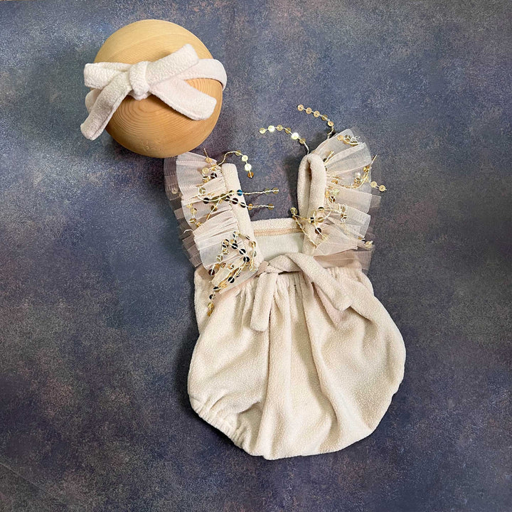A romper laid flat from the back, with a matching bow tie at the waist, tulle ruffle straps with sequins on both sides, a bow headband at the upper left.
