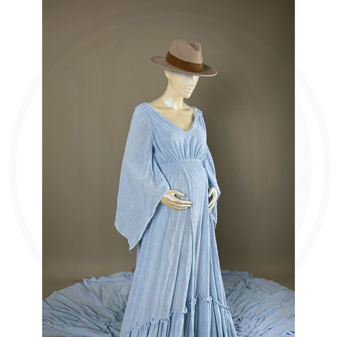 A  Daphne light blue long-sleeved V-neck gown is displayed on a mannequin, with hands positioned on the simulated belly. The dress features a high front slit and its hem and train are spread out on the floor.