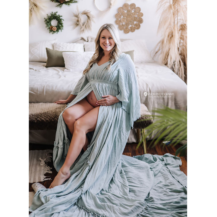 A woman sits on a bedroom bench wearing a Daphne pale smokey blue gown with the center open, revealing her belly. Her hands are on her belly, and the hem lies on the floor.