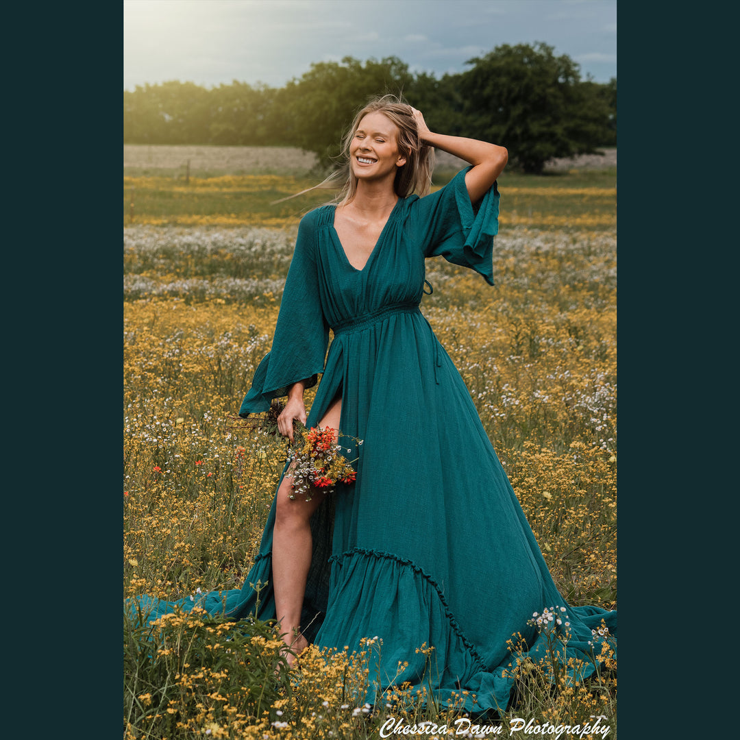 A woman in a Daphne emerald dress stands in a wildflower field, holding flowers in one hand and touching her hair with the other. The dress has a V-neck, flared sleeves, and a high slit.