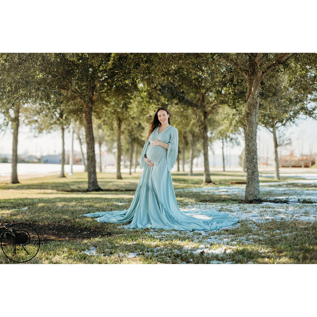 A pregnant woman in a light blue long-sleeve dress stands under trees on grass with snow, holding her belly.