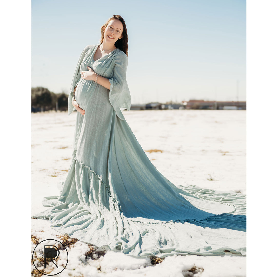 A pregnant woman in a Daphne light blue slit dress with a shawl stands on snow-covered grass, holding her belly.