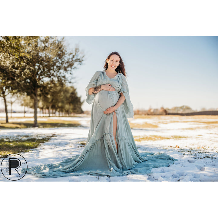 A pregnant woman in a Daphne light blue slit dress with a shawl stands on snow-covered grass, holding her belly.
