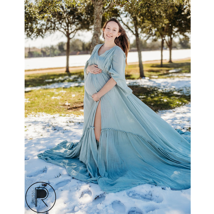 a pregnant woman in a light blue long-sleeve slit dress stands on snowy grass, hands on her belly.