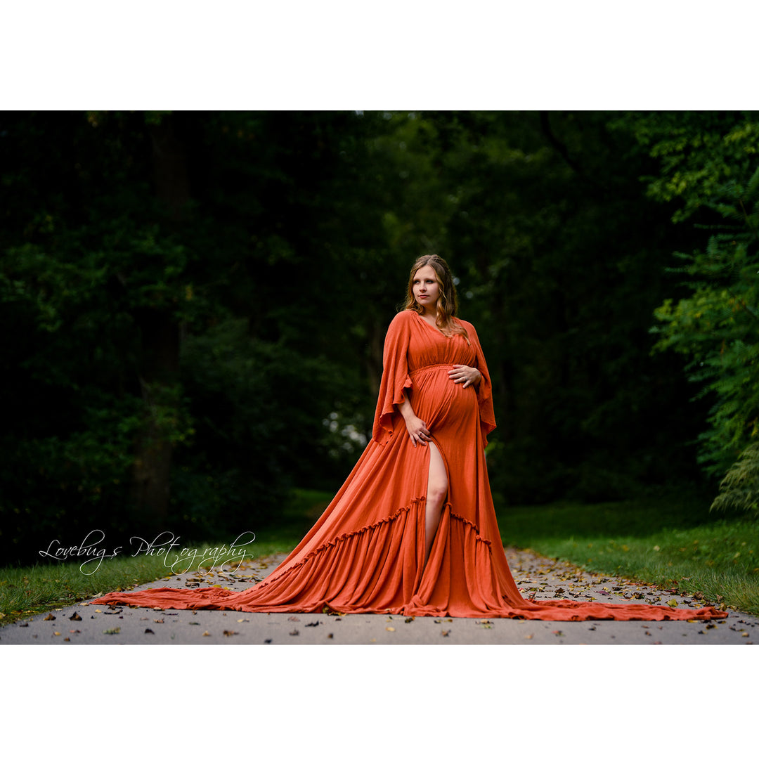 A pregnant woman in a Daphne rust red stands on a forest path with her skirt spread on the ground. She has one hand on her belly, and the dress features a high slit and cape sleeves.
