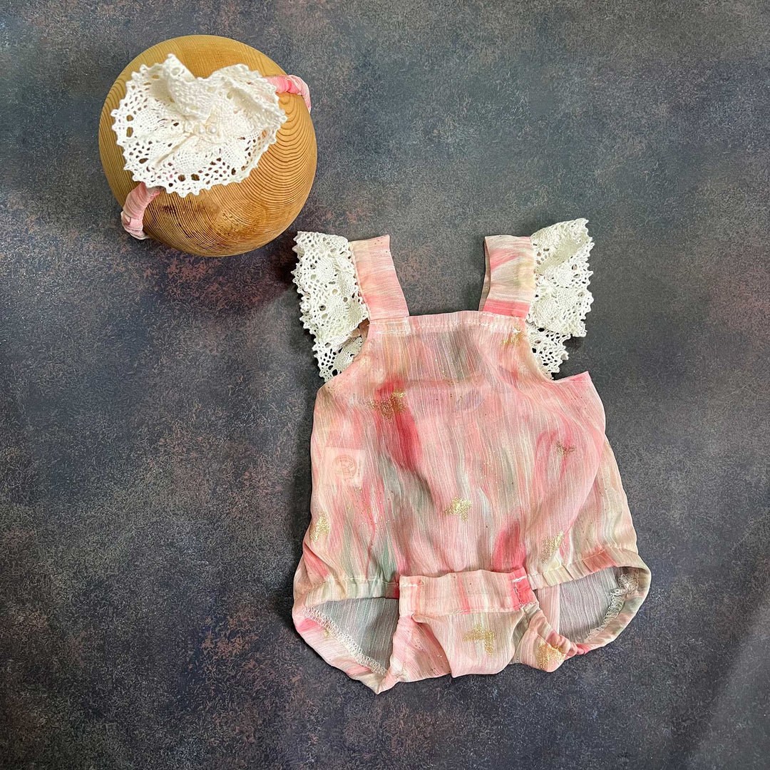 A pink tie-dye baby suspender romper laid flat, white crochet lace on both shoulder straps, a lace-flower headband at the upper left.