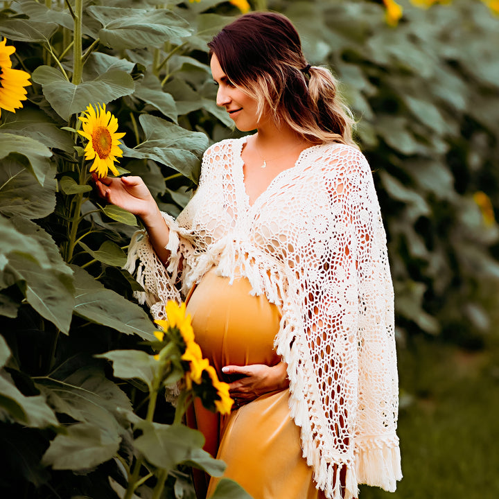 A pregnant woman in a KO-9 slip dress and a white crochet poncho stands in a sunflower field. She holds a sunflower with one hand and her belly with the other.
