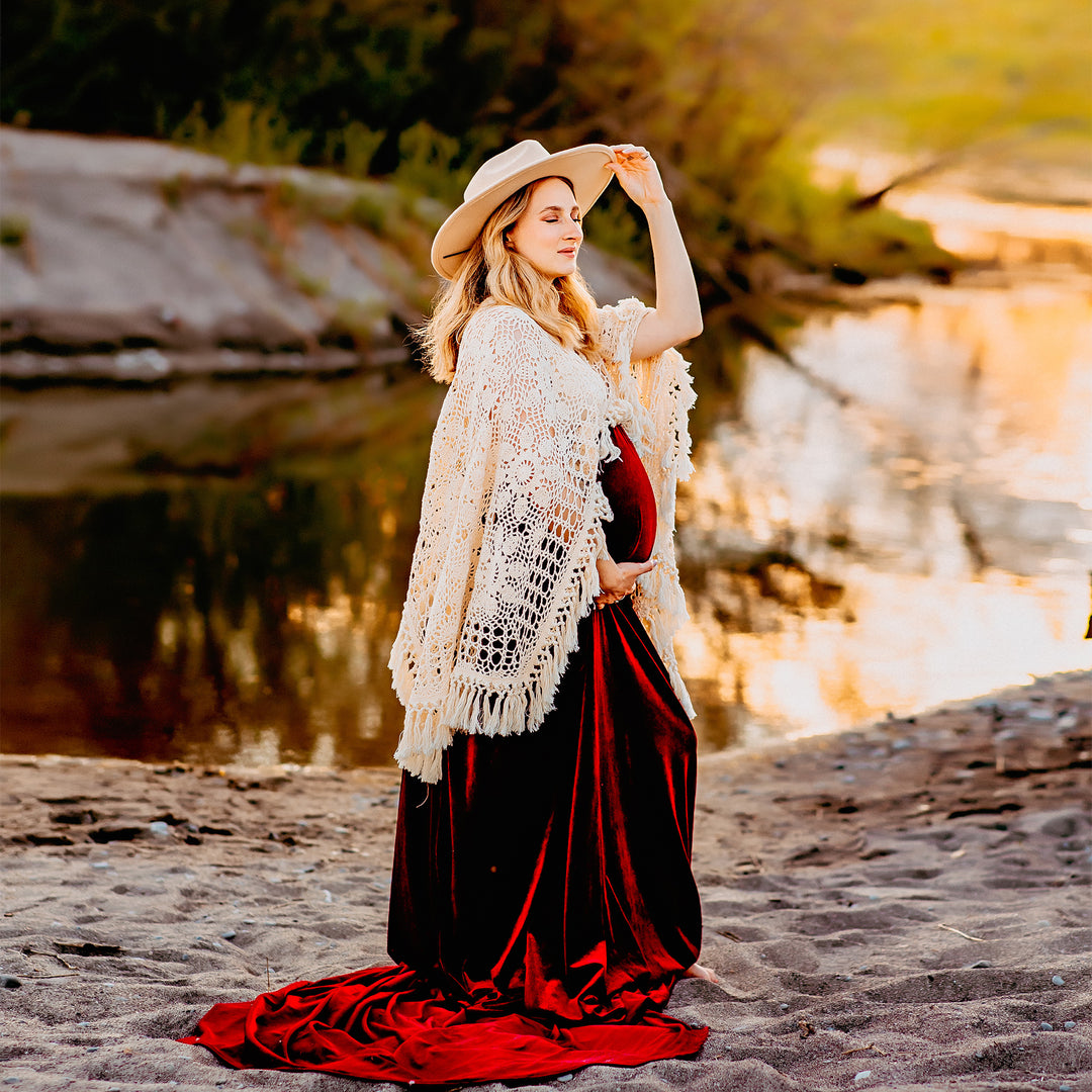 A pregnant woman wearing Edwina KO-14 velvet gown, a white crochet poncho, and a hat stands on a sandy ground near water. She holds her belly with one hand and touches her hat with the other.
