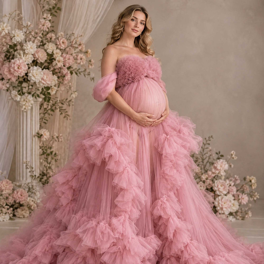 A pregnant woman stands in a pink strapless ruffled tulle gown, holding her belly; the dress features multi-layered ruffles.