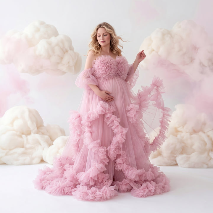 A pregnant woman in a pink off-shoulder ruffled gown holds her belly and lifts the skirt; the dress features multi-layered ruffles.